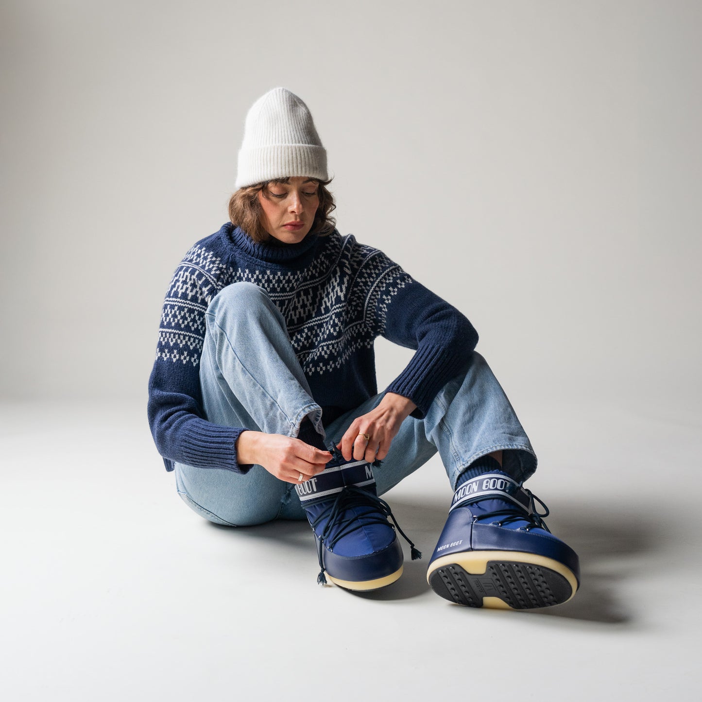 A person sits on the studio floor in light jeans and a white beanie, lacing up Moon Boot's Icon Low Nylon in blue.