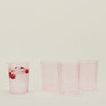 On a light background, four pink transparent Hawkins New York Essential Glassware cups (Set of 4) Large, three empty and one filled with water and raspberries, embody retro-inspired elegance with stackable borosilicate glass.