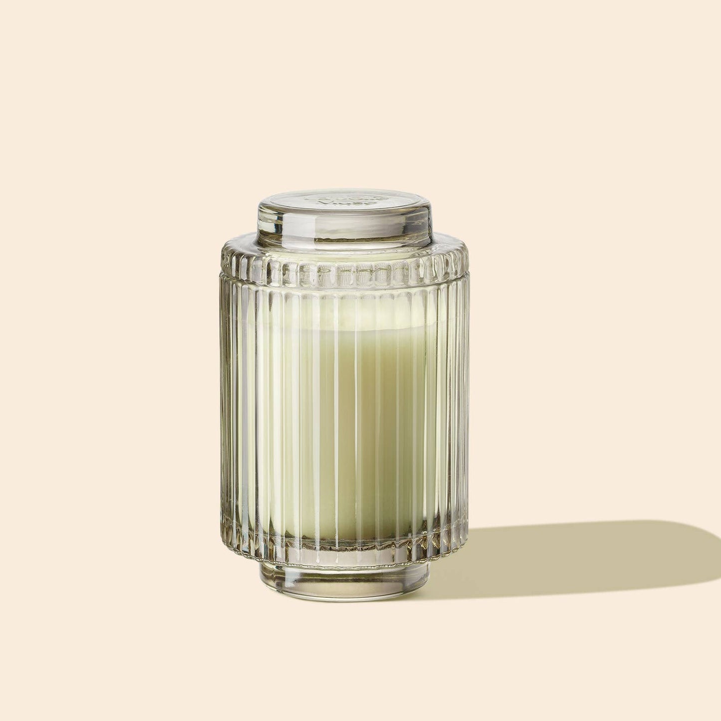 The Amelie - Kyoto Vine & Moss 7oz Candle by La Jolie Muse, showcasing a ribbed glass jar filled with cream-colored natural soy wax and topped with a matching lid, rests against a plain beige background, ready to transport you with its enchanting forest fragrance.