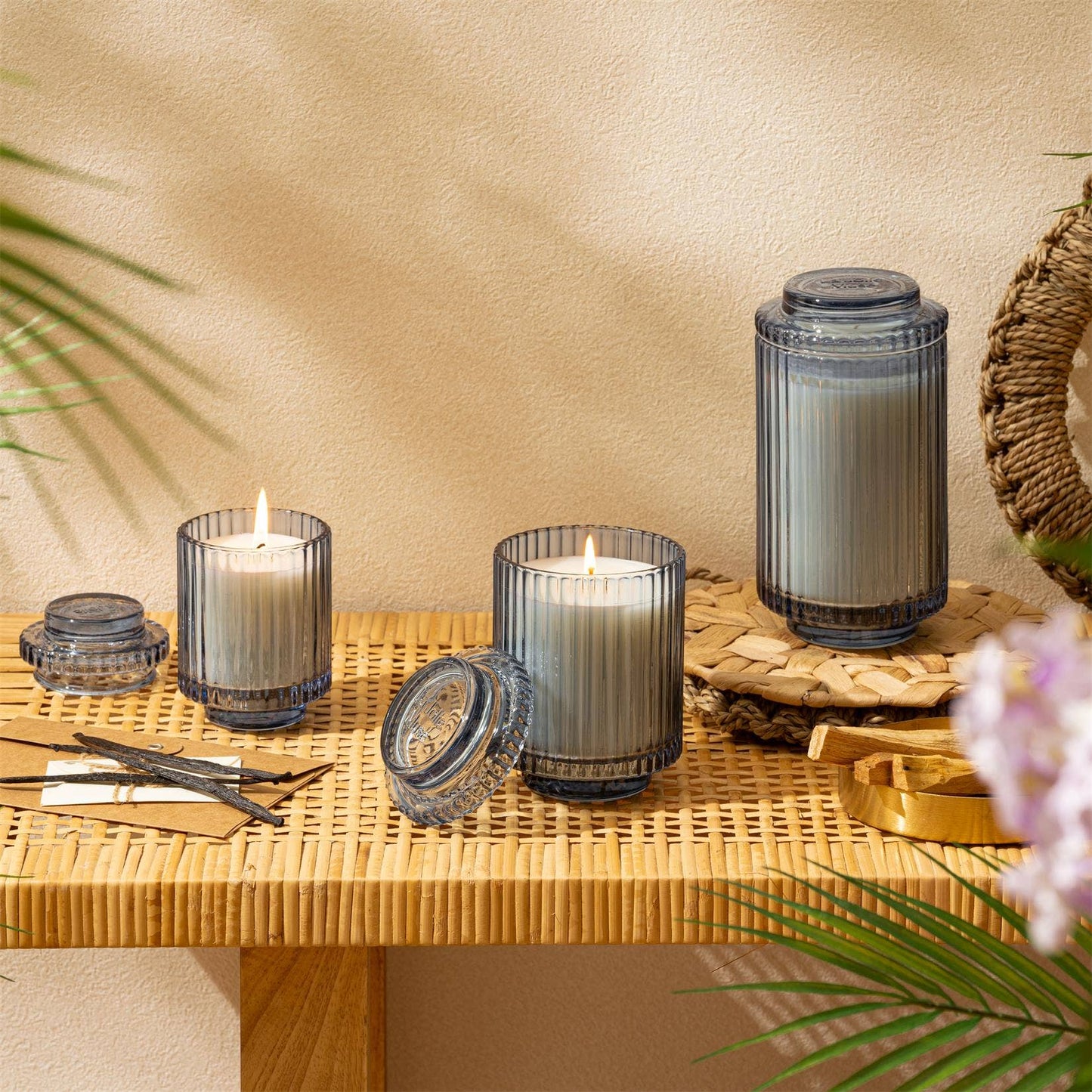 Three Amelie - Bora Bora 7oz candles by La Jolie Muse, encased in elegant keepsake containers, sit on a woven wooden table with decorative accents like a small leaf and a circular wicker piece against a beige wall.