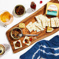 A wooden board with crackers, Ekone - Pantry Lemon Pepper Smoked Oysters, lemon wedges, and a jar of hot sauce. A glass of drink, a can, and a bottle top are nearby. A folded blue cloth lies beside the board.