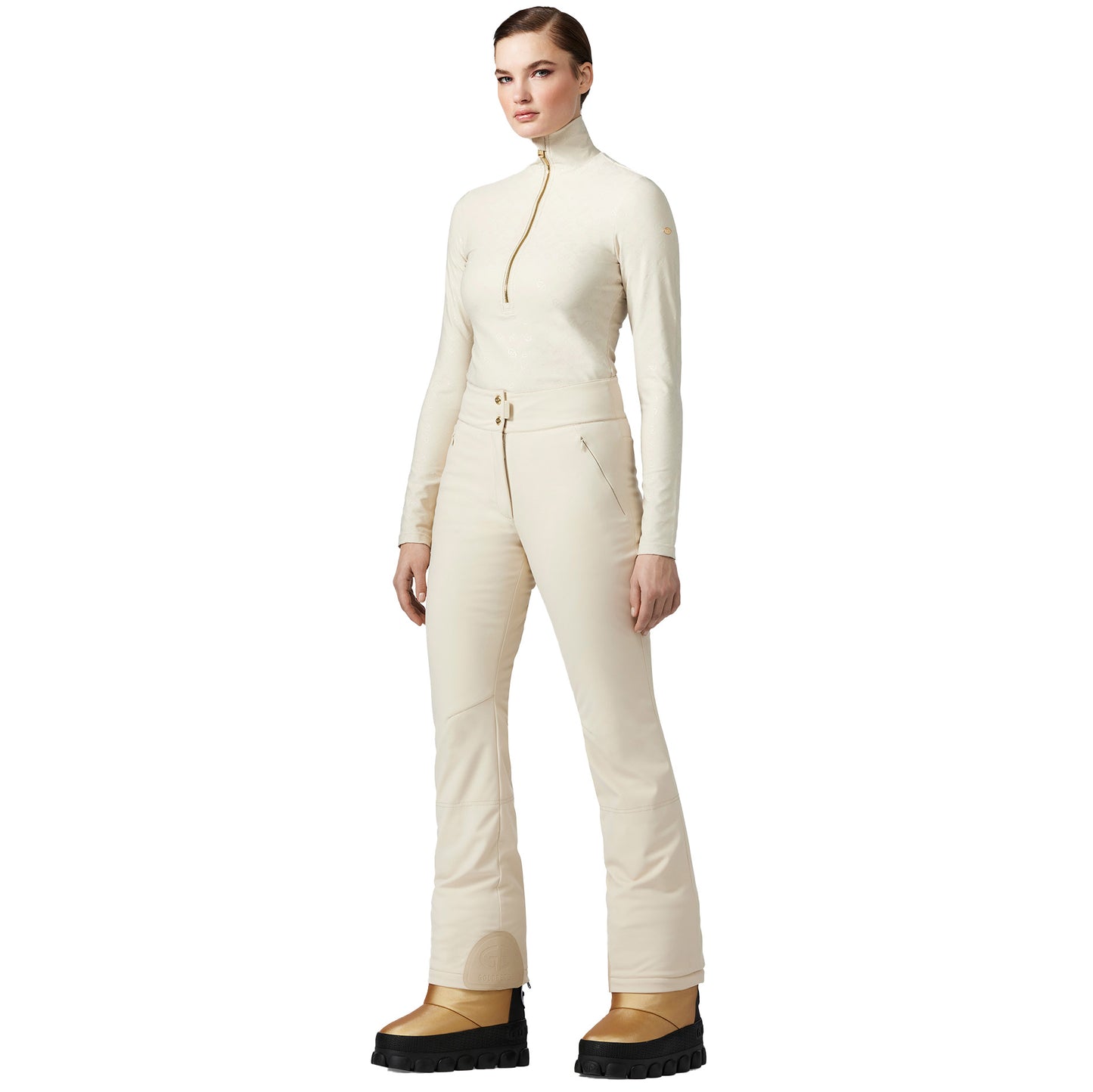 A person wearing Flair Ski Pants in champagne from the brand Goldbergh and brown boots stands against a plain white background.