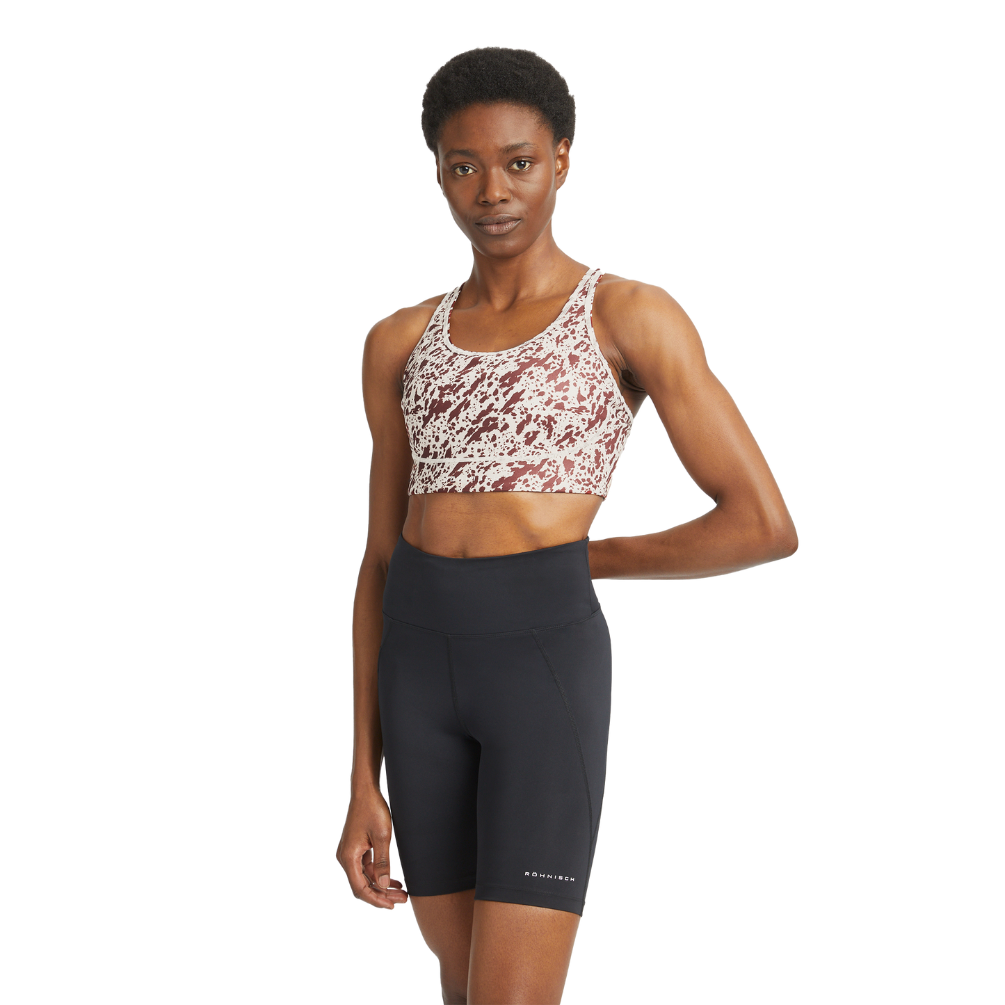 A person wearing the Flattering Sports Bra by Rohnisch, featuring a pattern and logo, pairs it with black high-waisted bike shorts while posing against a plain background.