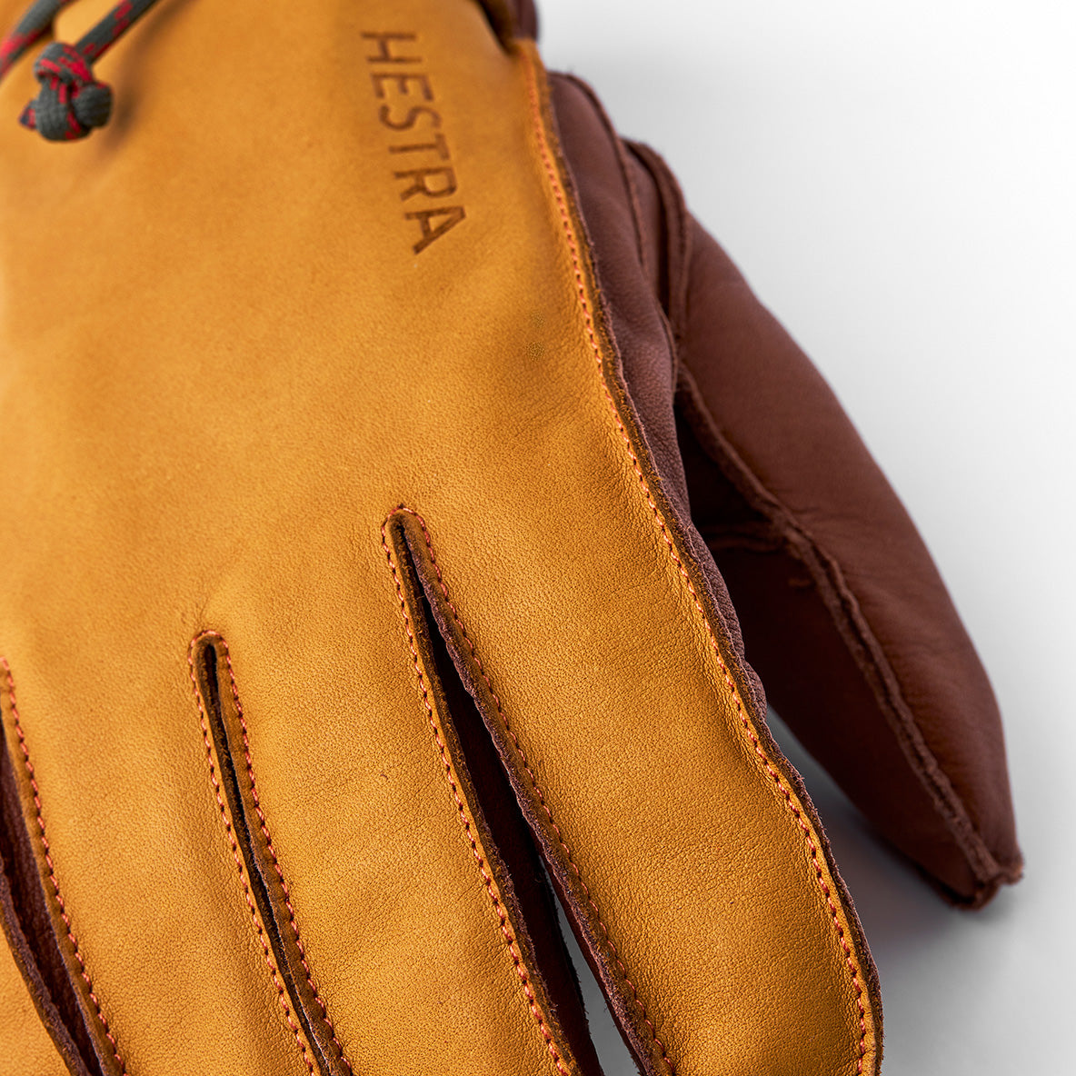 Close-up of a tan cowhide leather glove, "Wakayama, Cork/Brown" by Hestra, with embossed branding on the back. The ski glove shows visible stitching, brown finger trim, and has removable liners for added warmth and versatility.