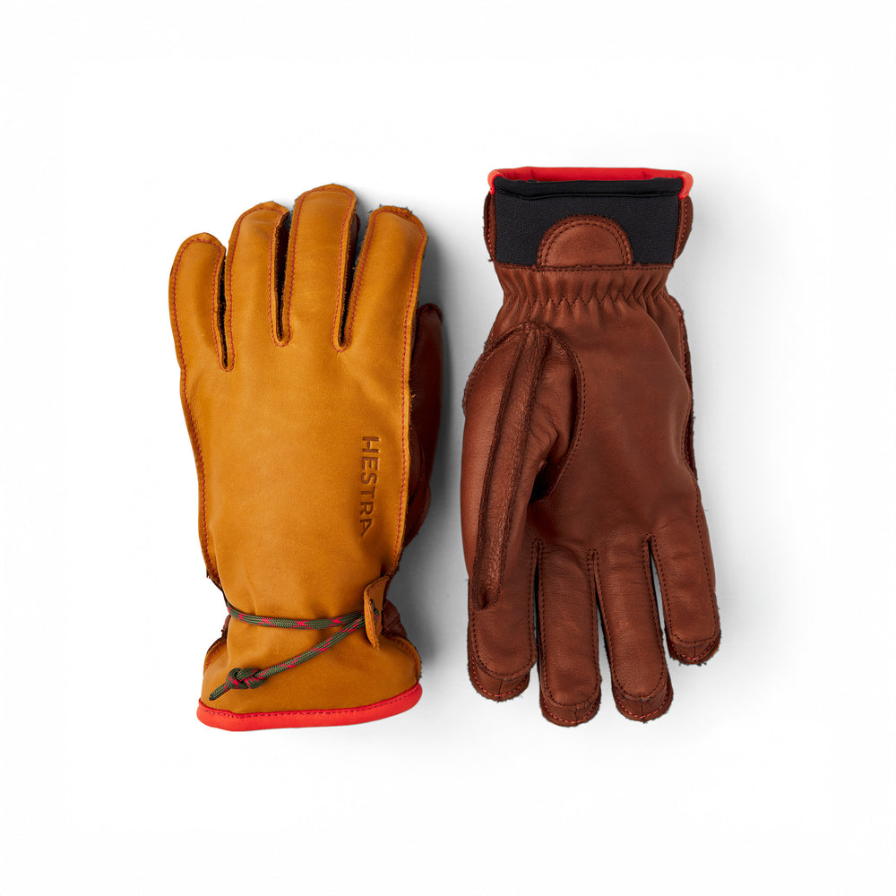 The Hestra Wakayama gloves in cork/brown are crafted from cowhide leather, featuring red trim and a branded logo on the left glove's palm-down, ski-glove design with a cinch cord. The right glove is shown palm-up. These gloves are perfect for the slopes with their removable liners.