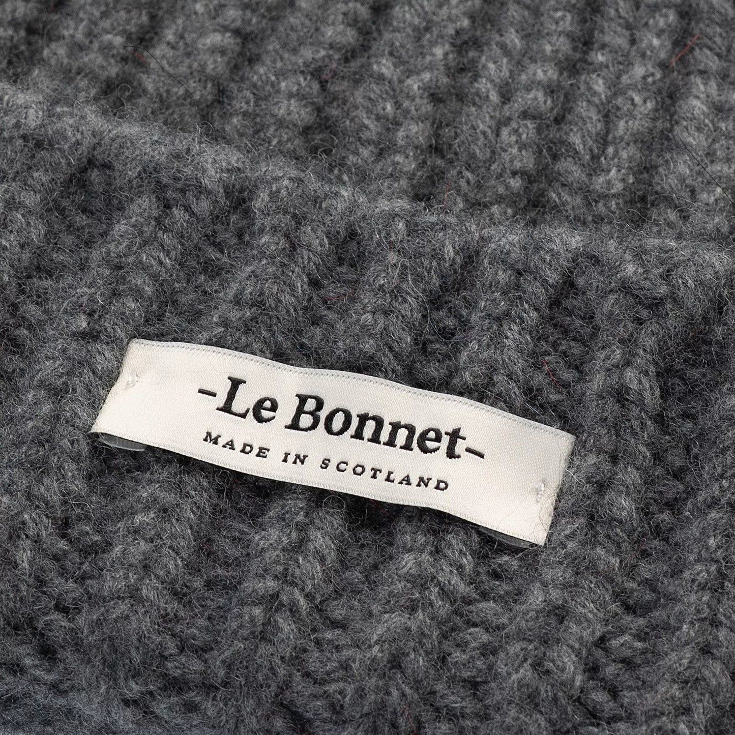 Close-up of grey slate knitted lambswool fabric with a white label reading "Le Bonnet Made in Scotland," featuring the unique Le Bonnet logo. Product: Logo Beanie.