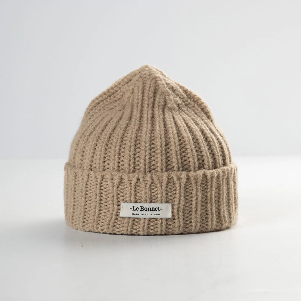 The Logo Beanie in Sand by Le Bonnet is a beige ribbed knit crafted from soft lambswool, featuring a folded cuff and a small logo on a plain white background.