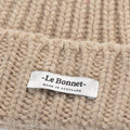 A close-up of the Logo Beanie in Sand showcases its beige knitted lambswool with visible stitching. The Le Bonnet logo tag reads "Made in Scotland.