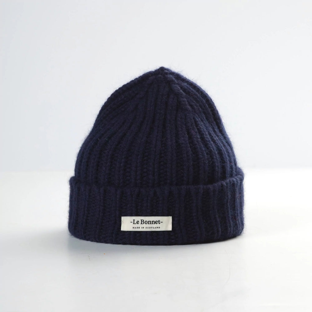 The Le Bonnet Logo Beanie, Midnight, is a dark blue ribbed knit with a folded brim showcasing the brand logo. Crafted in Scotland, this durable beanie provides style and functionality against a light background.