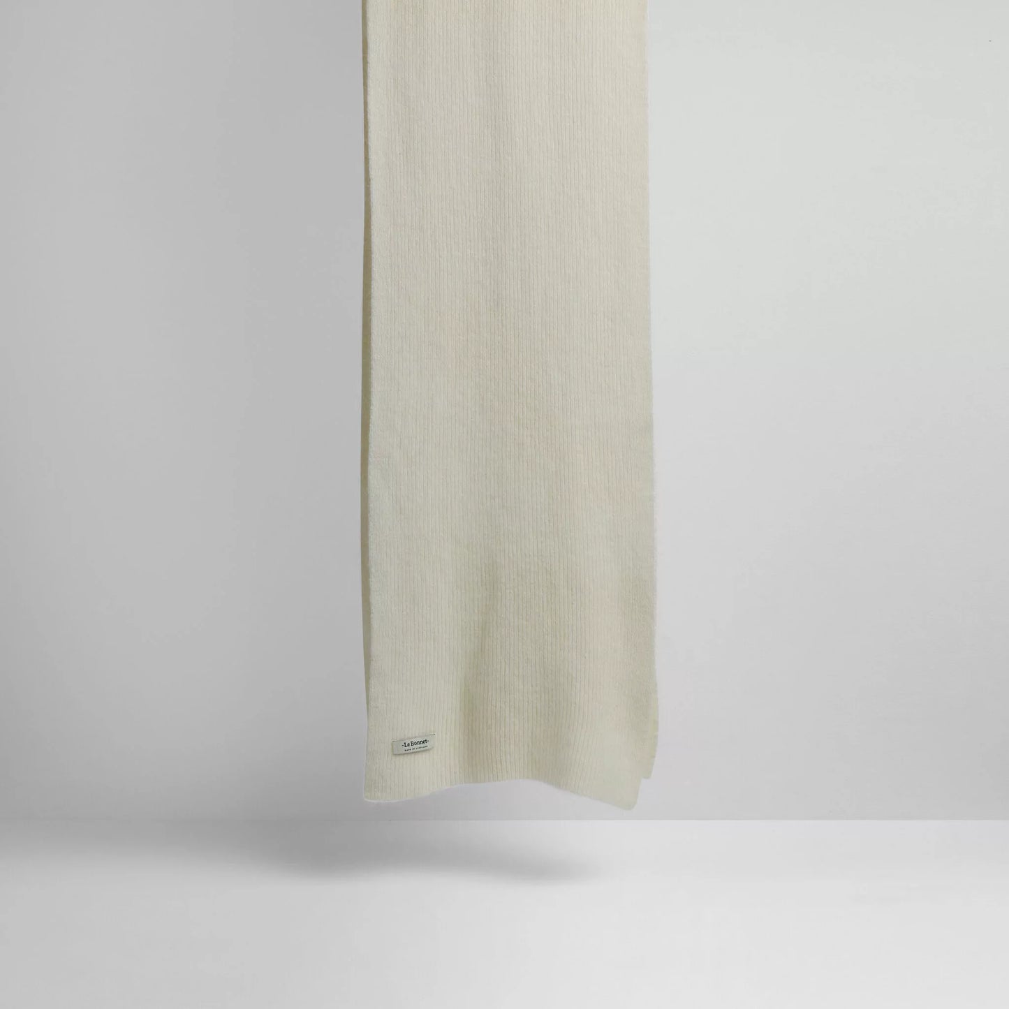 A plain, light-colored Le Bonnet Scarves, Snow lambswool scarf hangs against a neutral background highlighting its biodegradable properties with a small tag near the edge.