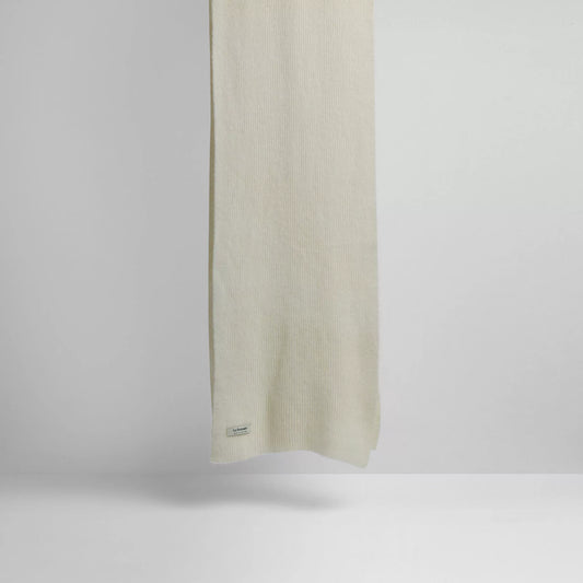 A plain, light-colored Le Bonnet Scarves, Snow lambswool scarf hangs against a neutral background highlighting its biodegradable properties with a small tag near the edge.