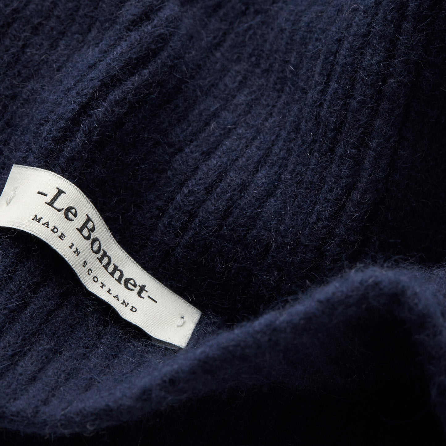 Close-up of a dark blue lambswool knit fabric with a sewn white label reading "Le Bonnet" and showing the product name "Scarves, Midnight," indicating it is made in Scotland.