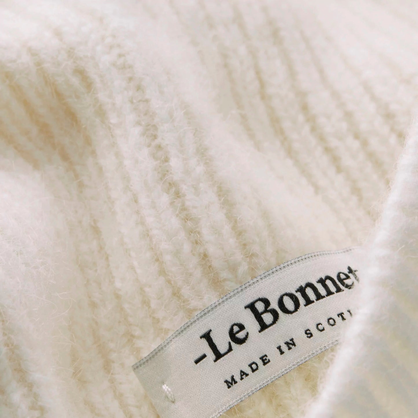 A close-up of cream-colored knitted lambswool fabric resembling a cozy scarf, featuring a "Le Bonnet, Made in Scotland" label. Product Name: Scarves, Snow.