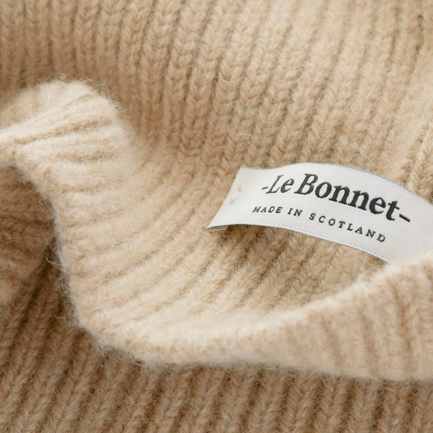 A close-up of the soft, biodegradable lambswool fabric in "Scarves, Sand" by Le Bonnet features a label stating "Le Bonnet - Made in Scotland.