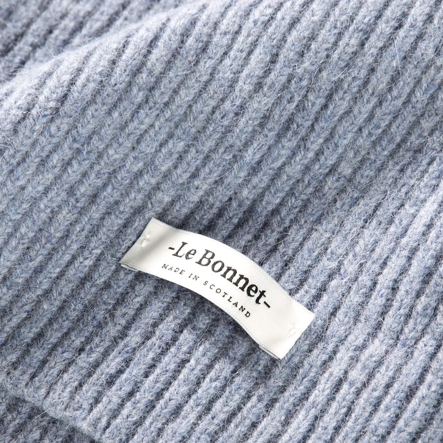 A close-up of a light gray lambswool knitted fabric shows a white label with "Le Bonnet Made in Scotland" written in black.