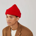 Wearing a Le Bonnet Crimson Balaclava and a brown coat over a white, limited edition Merino wool sweater, the person gazes thoughtfully to the side against a plain backdrop.