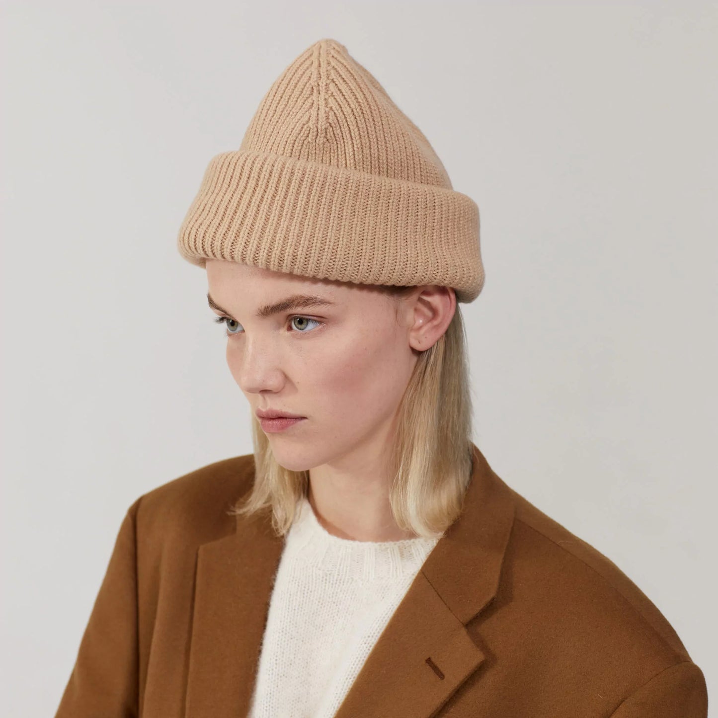 A person is wearing the limited edition Le Bonnet Balaclava in Sand, paired with a brown coat and white sweater, against a plain background.