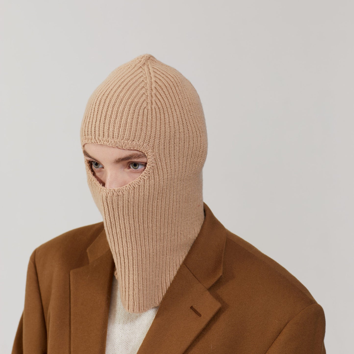 Someone wearing a limited edition Sand Balaclava by Le Bonnet, made from cashmere, paired with a brown coat, gazes off to the side.