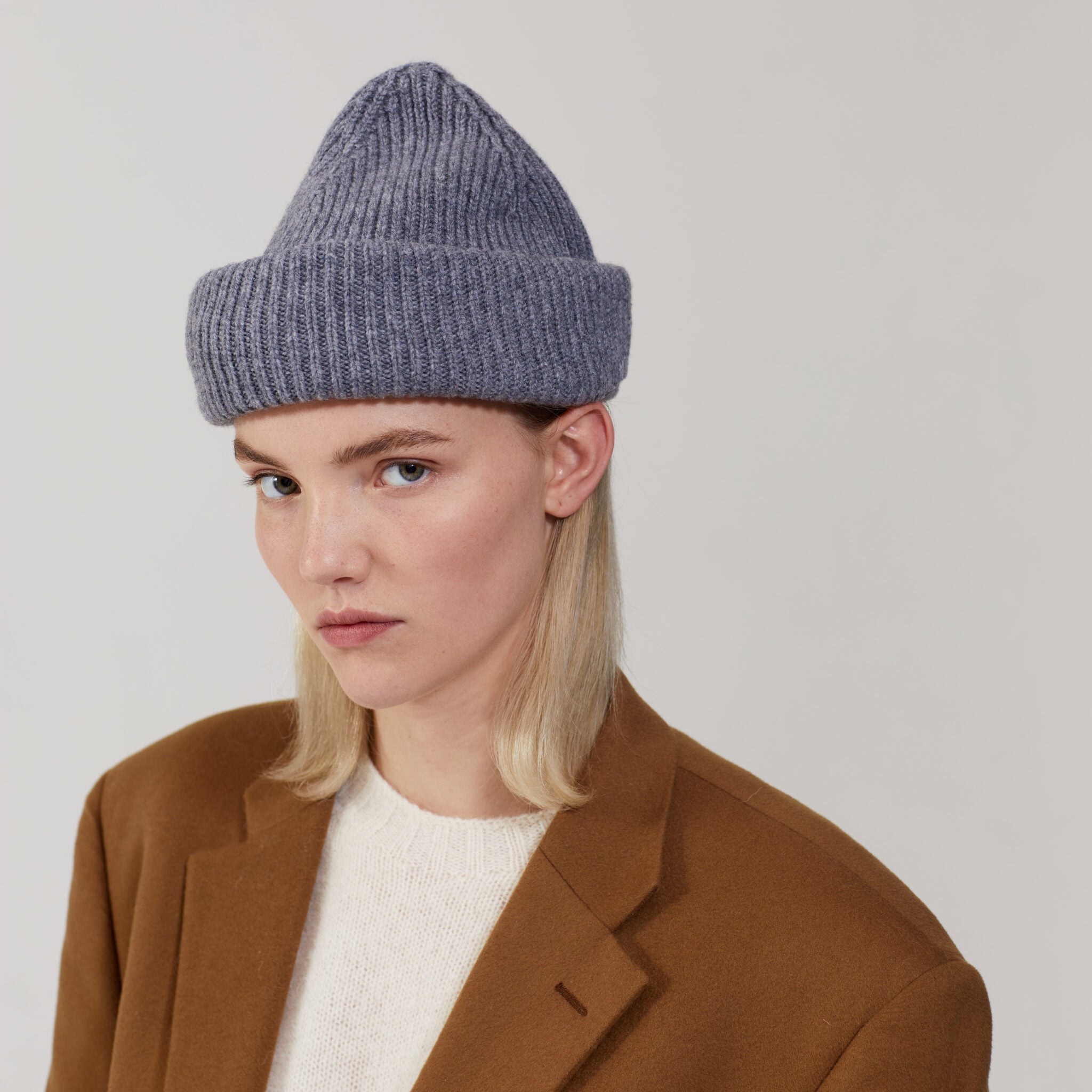 Dressed in a limited edition brown coat and cozy white sweater, a person with long blond hair completes the look with Westerlind's Smoke Balaclava, made of luxurious Merino wool, all set against a plain background.