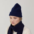 A person in a cream sweater, gazing downward against a plain background, is accessorized with Le Bonnet's Midnight scarves and a navy blue lambswool knit hat.