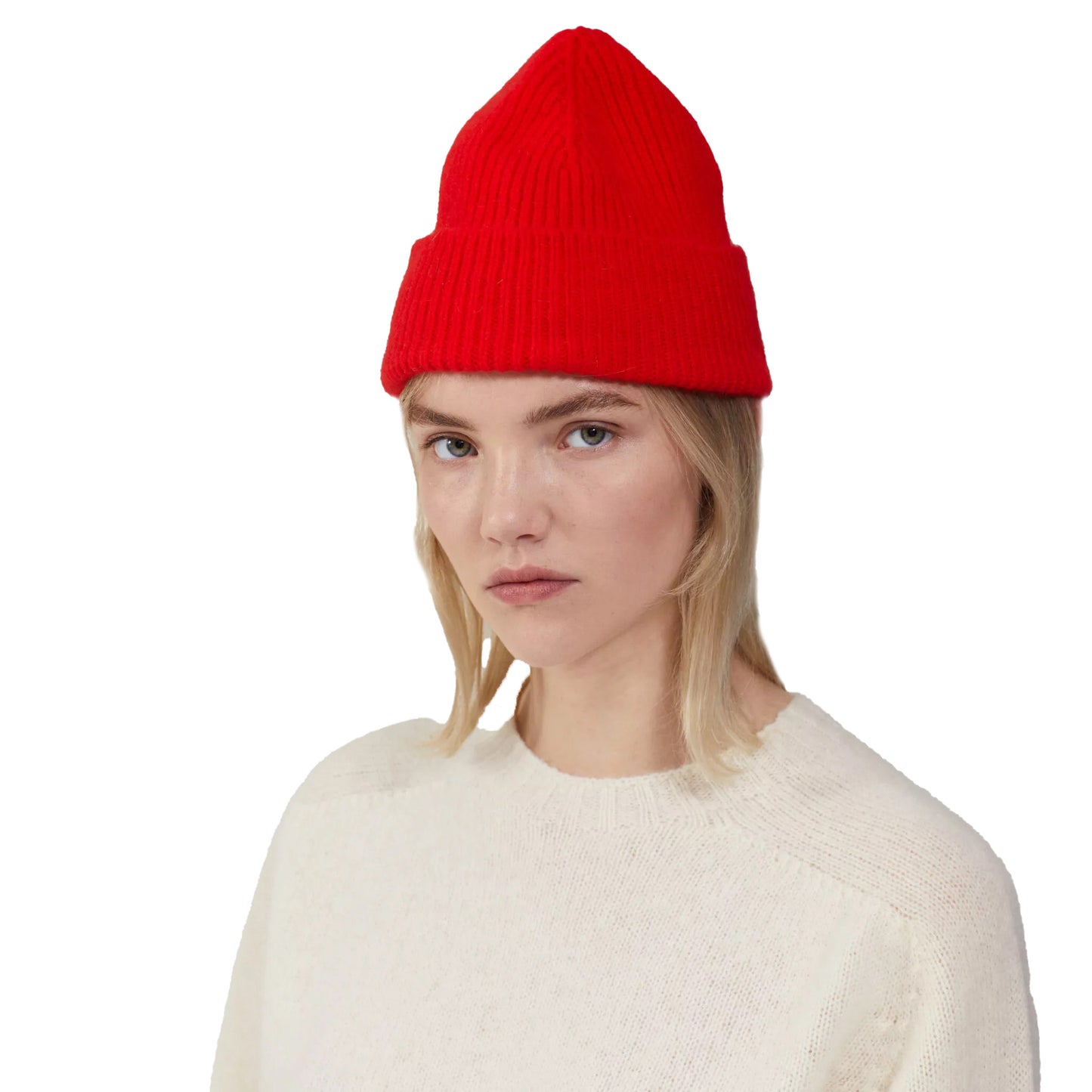 A person with long blonde hair, sporting a red wool Beanie from Le Bonnet and a white sweater, gazes at the camera against a simple backdrop, radiating cozy vibes akin to those of a crisp day in Scotland.