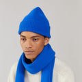 A person wears a bright blue knit hat and a soft Le Bonnet Royal Azure lambswool scarf, gracefully draped over a light sweater, gazing thoughtfully to the side.