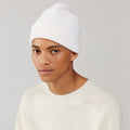 Wearing a Snow Beanie by Le Bonnet and a cream sweater, a person gazes slightly to the side against a light gray backdrop, exuding cozy Scottish charm.