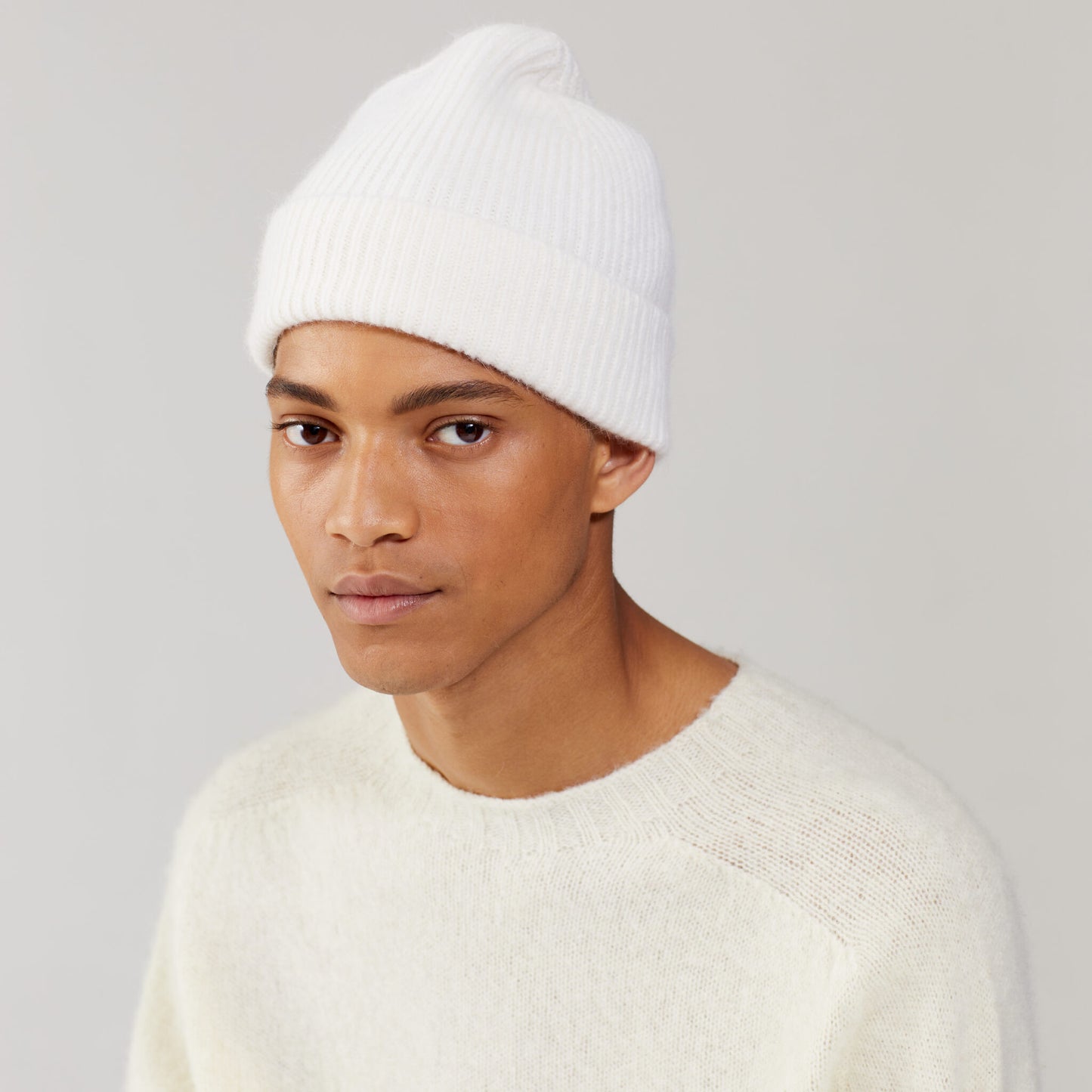 Wearing a Snow Beanie by Le Bonnet and a cream sweater, a person gazes slightly to the side against a light gray backdrop, exuding cozy Scottish charm.