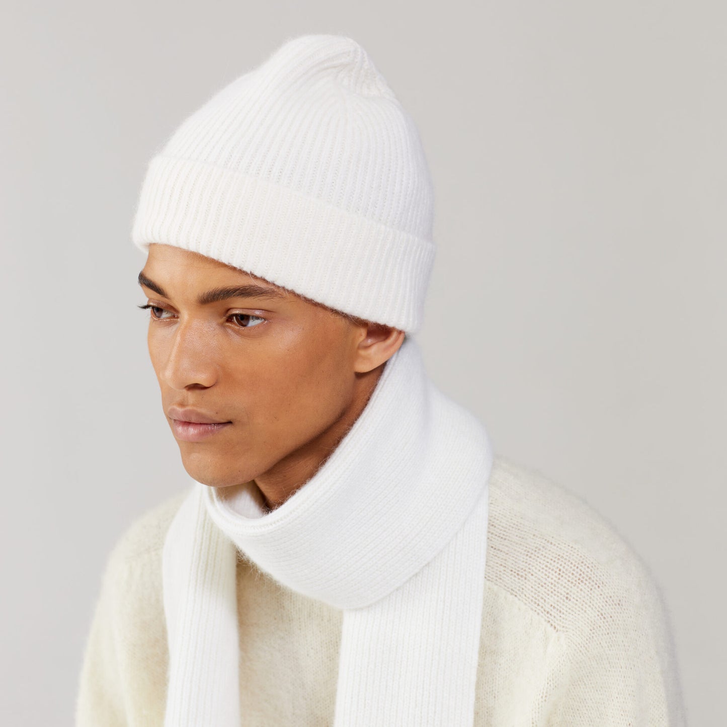 A person dons a white knit beanie and Le Bonnet's cozy Snow scarf, paired with a matching sweater against a plain background.