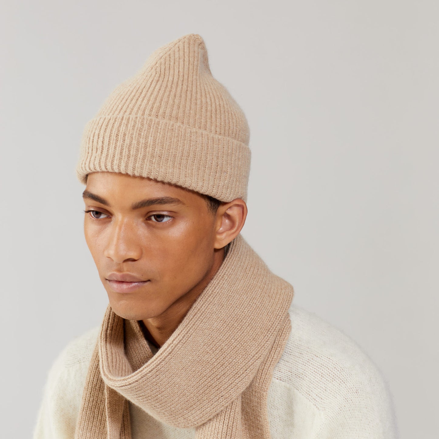 A person wearing a matching beige Le Bonnet lambswool knit hat and Sand scarf stands, expression neutral, in a light cream sweater against a plain background.