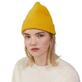 A person with blonde hair is wearing a yellow Beanie by Le Bonnet and a cream-colored sweater, looking straight ahead.