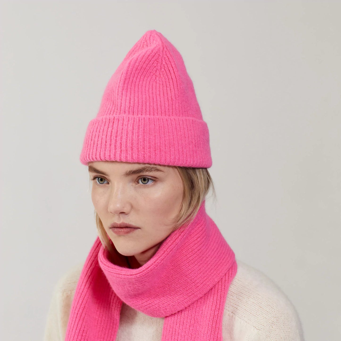 A person wears a bright Bubblegum lambswool knit hat and matching Le Bonnet scarf, facing slightly to the side with a neutral expression against a plain background.