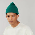 A person wearing a Le Bonnet "Beanie, Pine" in green and a white sweater stands against a plain background.