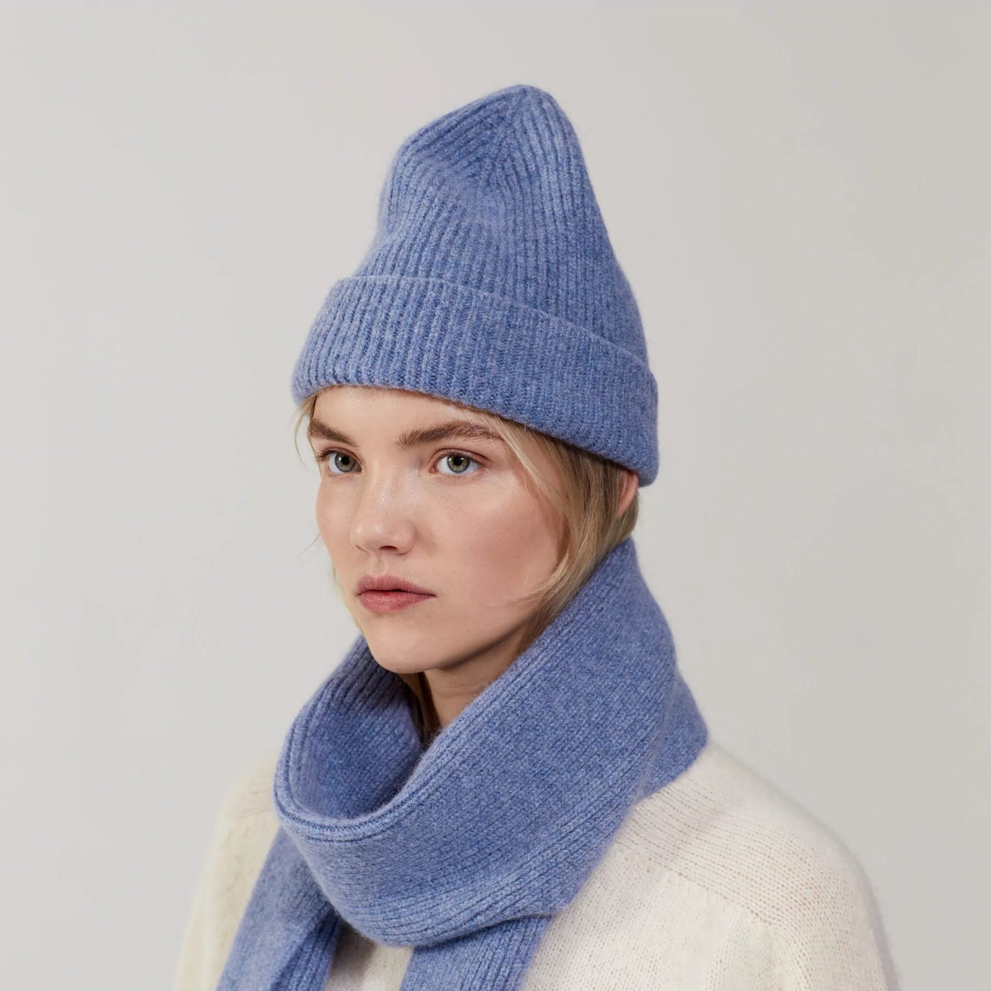 A person wearing a blue Le Bonnet knit hat and a matching Washed Denim scarf, gazes to the side against a plain backdrop.