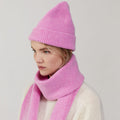 In a plain setting, a person wears Le Bonnet's pink knit hat, the Taffy scarf, and a cream sweater, glancing sideways.