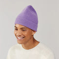 A person in a Lavender Beanie by Le Bonnet and a cream sweater of biodegradable fabric smiles, glancing sideways.