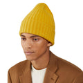 A person wearing a yellow Large Beanie by Le Bonnet, paired with a white sweater and brown coat, exudes extreme comfort as they look to the side against a plain white background.