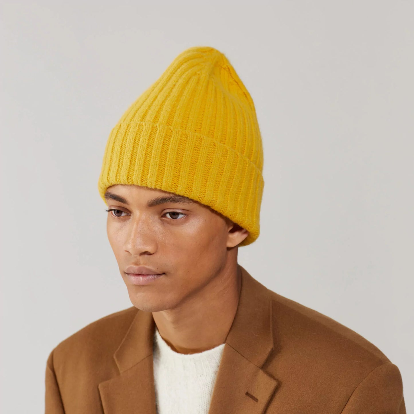 A person wearing Le Bonnet's Large Beanie in Mustard with a brown coat is looking away from the camera against a neutral background.