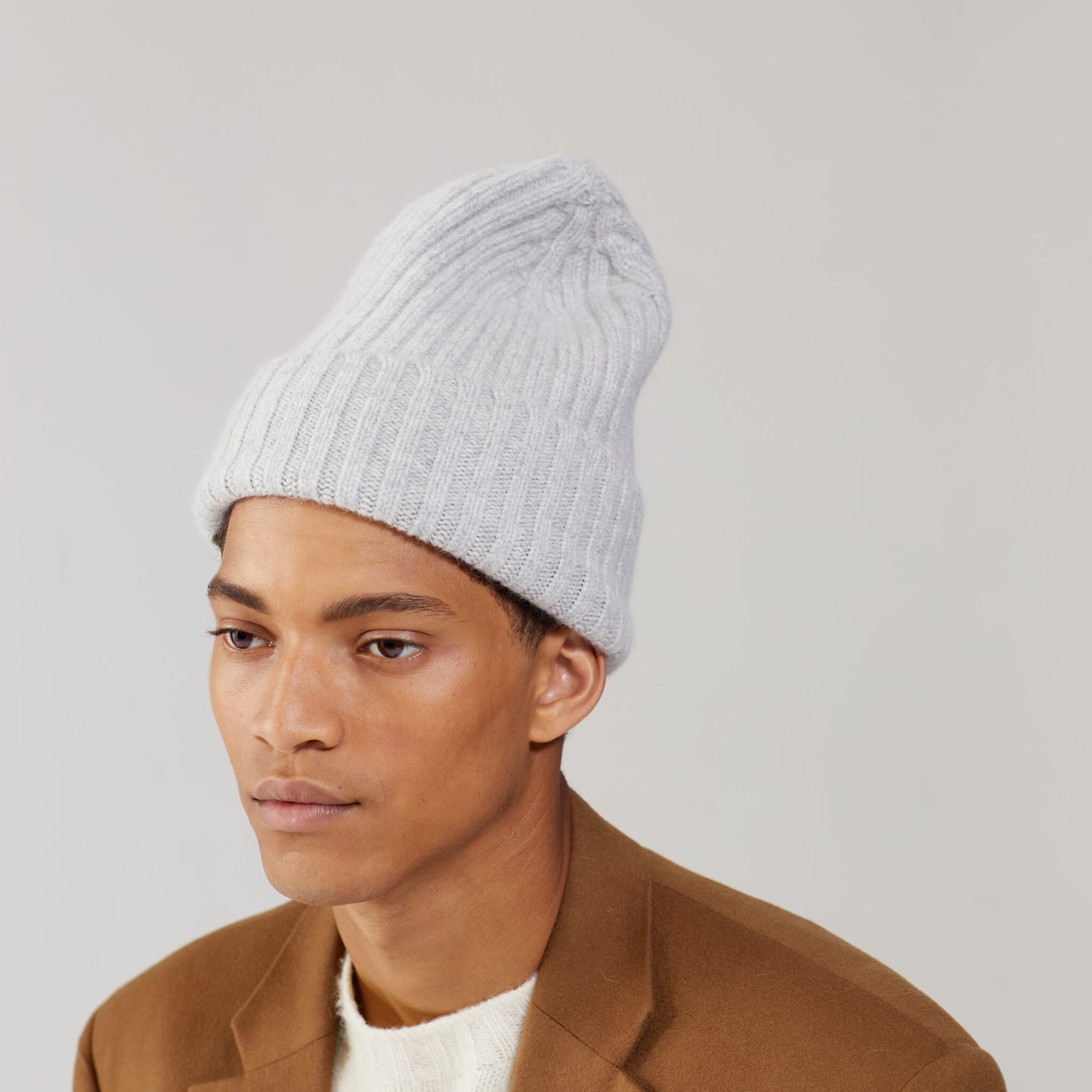 The person wears a Large Beanie in Silver by Le Bonnet, possibly a chunkier version made in Scotland, paired with a brown jacket against a plain background.