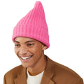 A person wearing a large pink Le Bonnet lambswool knit beanie and a brown jacket smiles warmly against a plain background.