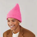 A person smiles warmly against a plain background, exuding extreme comfort while wearing a Bubblegum Large Beanie by Le Bonnet and a brown coat.