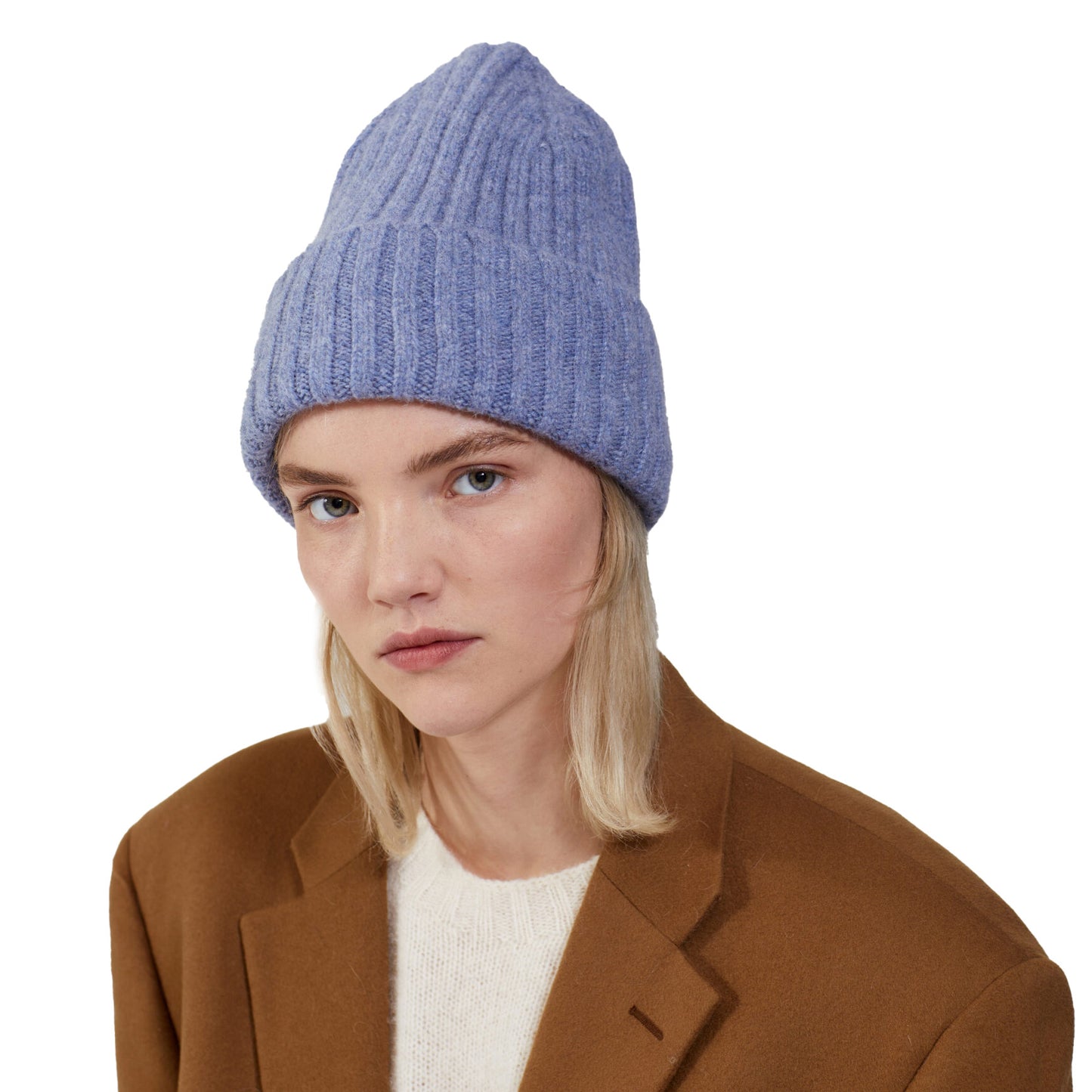 A person exudes comfort in a chunky blue Le Bonnet Large Beanie, paired with a cozy white sweater and a sturdy brown coat against a plain background.