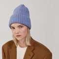 A person wearing Le Bonnet's Large Beanie in Washed Denim, a white sweater, and a brown coat stands against a plain background.