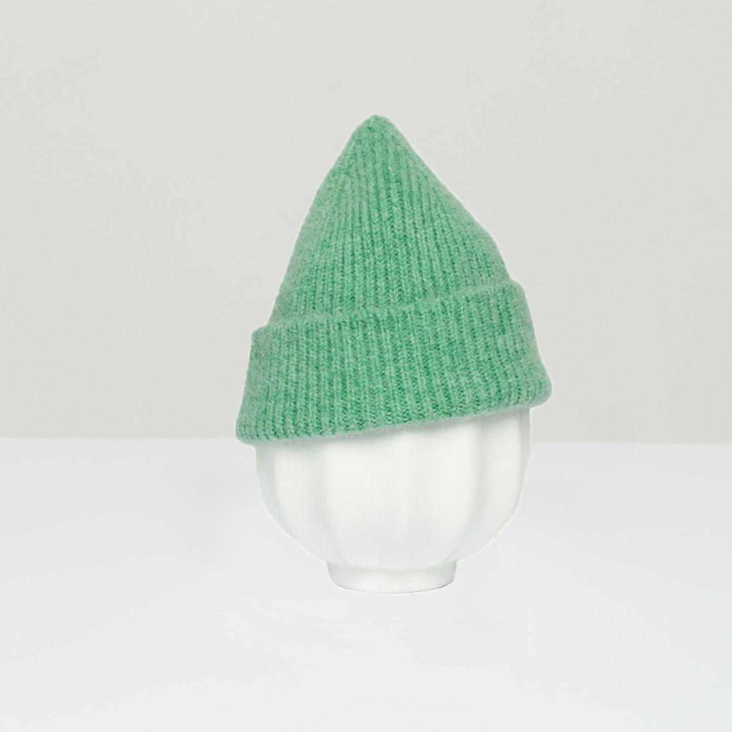 The Le Bonnet K Beanie in Jade elegantly sits on a white ceramic bowl against a simple background, highlighting its versatile one-size-fits-all design.