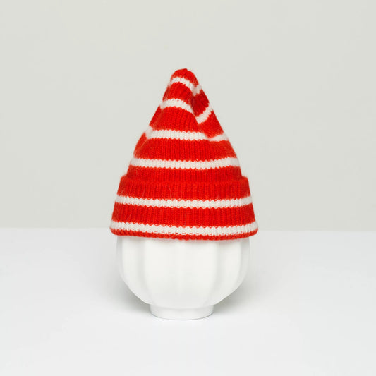 A white ceramic vase with a ribbed texture is topped with Le Bonnet's K Beanie, Crimson Stripe, a red and white striped knit hat made from soft Caregora for a cozy touch.