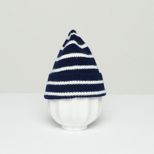 A white vase wearing Le Bonnet's K Beanie in Midnight Stripe, crafted from soft lambswool, takes on a conical shape. It rests elegantly on a white surface against a light gray background.