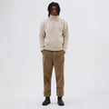 A person is standing against a plain white background wearing Margaret Howell's M's T Shape Roll Neck sweater in beige, paired with brown pants and black boots.