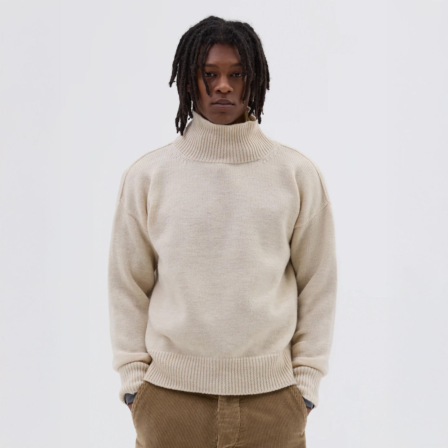 A person with dreadlocks wearing Margaret Howell's M's T Shape Roll Neck sweater in beige and brown pants stands against a plain white background.