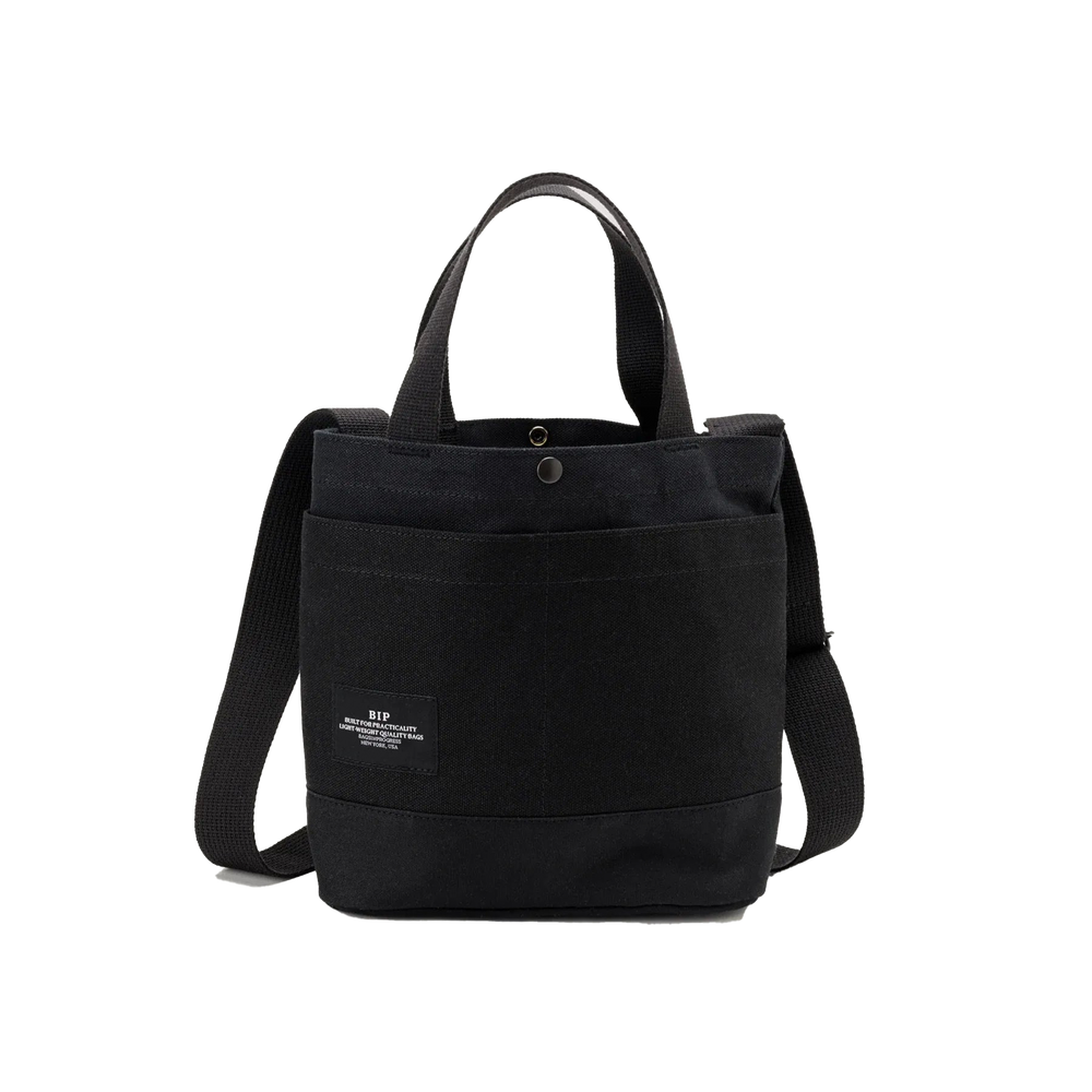 The New York Tote Small by Bags in Progress is a black cotton canvas tote bag featuring dual handles, an adjustable shoulder strap, and a front pocket with a small label on the front.