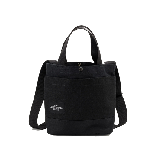 The New York Tote Small by Bags in Progress is a black cotton canvas tote bag featuring dual handles, an adjustable shoulder strap, and a front pocket with a small label on the front.